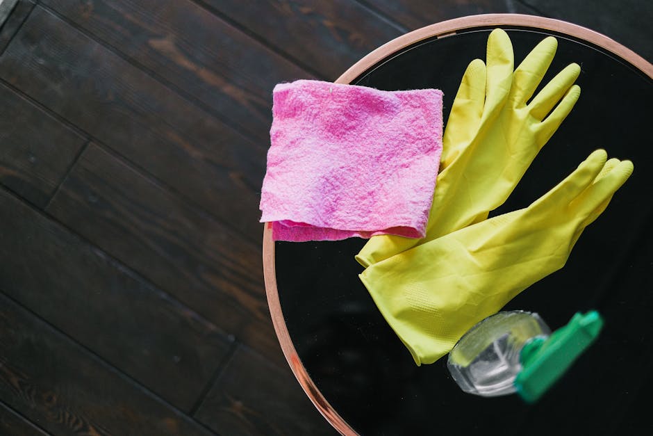 On a black circular surface, there is a pair of bright yellow rubber cleaning gloves, a pink microfiber cloth, and a partially visible spray bottle with a green nozzle. The surface is clean and shiny,