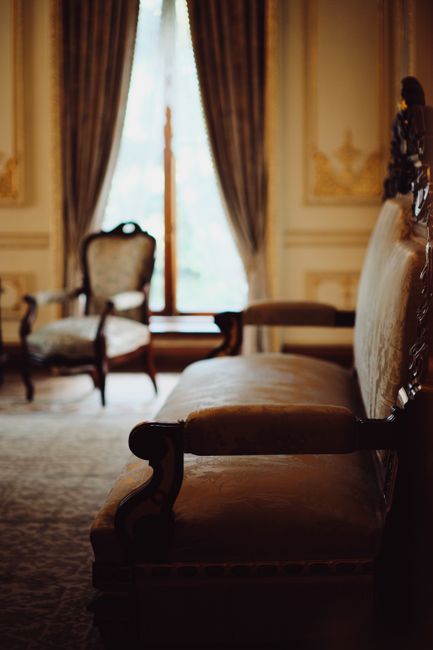 A vintage-style living room featuring a large, cream-colored, velvet-upholstered sofa with multiple matching cushions positioned in front of a window with heavy dark curtains tied back, allowing natur