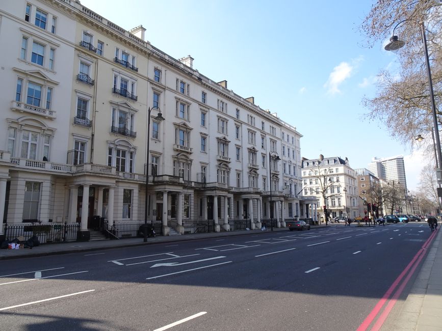 A city street scene in Pimlico Road, featuring a wide sidewalk with a black wrought iron fence bordering Victorian-style brick residential buildings. The sidewalk is clean and well-maintained, with a 